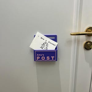 May include: A purple wall-mounted plastic mail holder featuring the text Amy's POST in light pink lettering. A white note is tucked inside, displaying a hand-drawn dinosaur illustration and the handwritten text I drew a dinosaur!