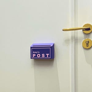 May include: A small, rectangular purple wall-mounted mail holder featuring a light pink border and the text "Amy's POST" in light pink lettering. The container has a horizontal slot at the top for inserting letters and is attached to a white door.