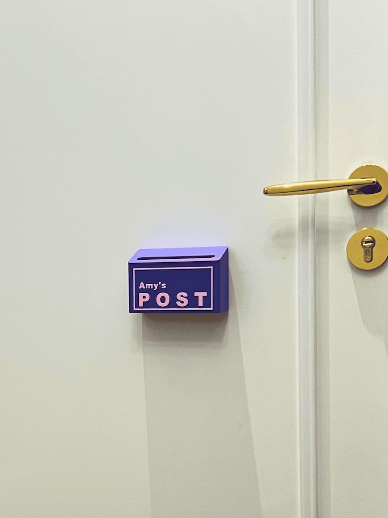 May include: A small, rectangular purple wall-mounted mail holder featuring a light pink border and the text "Amy's POST" in light pink lettering. The container has a horizontal slot at the top for inserting letters and is attached to a white door.
