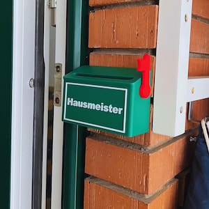 May include: A small green mailbox with a white border and the word "Hausmeister" printed in white on the front. The mailbox features a bright red flag attached to the right side, designed to signal when mail has been delivered or collected.