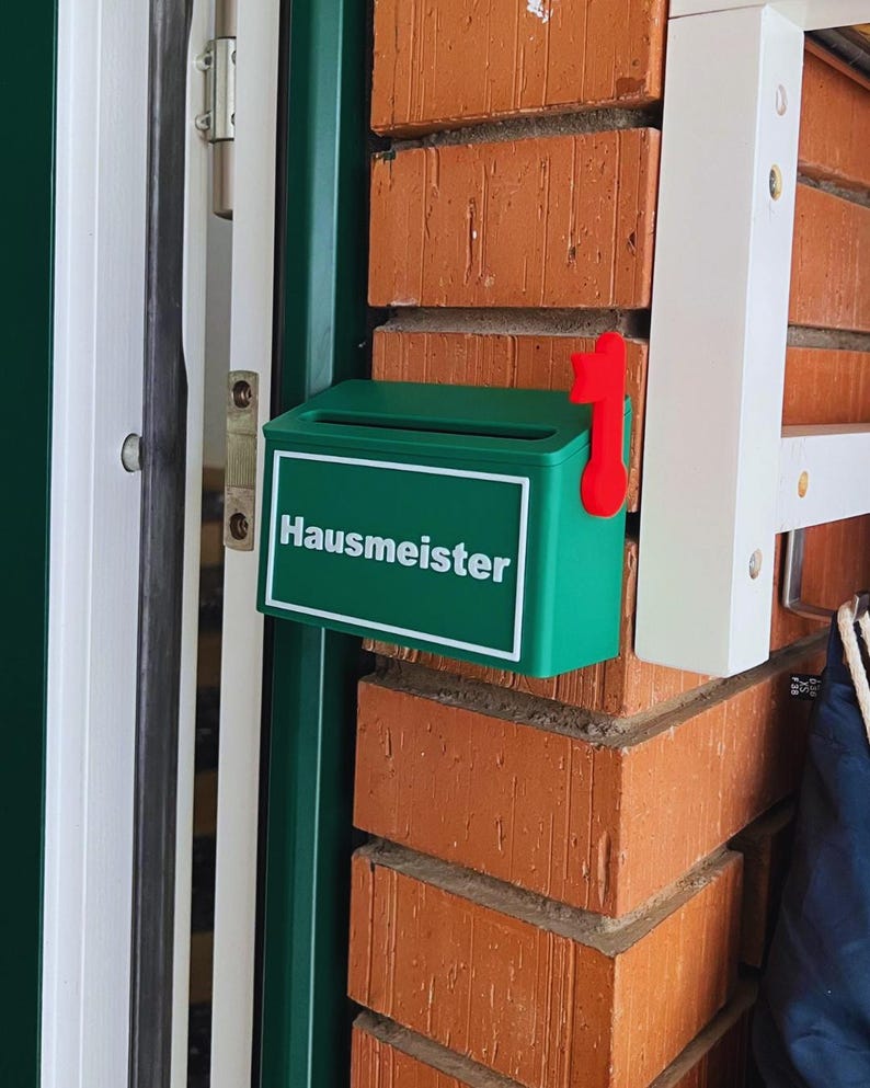 May include: A small green mailbox with a white border and the word "Hausmeister" printed in white on the front. The mailbox features a bright red flag attached to the right side, designed to signal when mail has been delivered or collected.