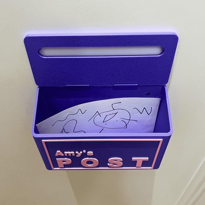 May include: A wall-mounted purple mailbox with a hinged lid and a rectangular opening. The front features a light pink border and the raised white text "Amy's POST". Inside the open compartment sits a white piece of paper with black ink drawings.