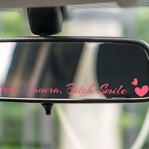 May include: A car rearview mirror with pink text that reads "Lights, Camera, Bitch Smile" and two pink hearts. The mirror is black and reflects the interior of the car.