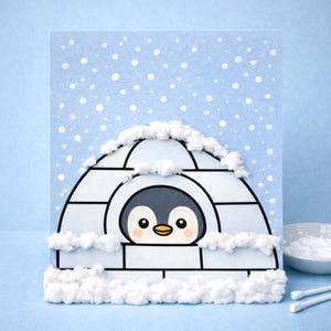 May include: A handmade greeting card featuring a cartoon penguin inside an igloo. The igloo is white with black outlines, and the penguin is gray and white. Fluffy white cotton simulates snow around the igloo and falling from the sky. The background is light blue.