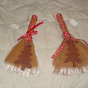 May include: Two decorative brooms with natural straw bristles and wooden handles. Each broom is adorned with a red ribbon featuring white snowflakes and a metal Christmas tree ornament. The tree ornament has the text "Merry Christmas".