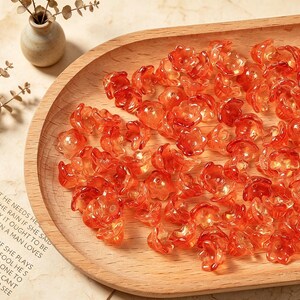 May include: A collection of small, translucent orange flower-shaped beads. Each bead features a five-petal design with a central hole for stringing. The beads are scattered across a smooth, light-colored wooden tray with a curved edge.