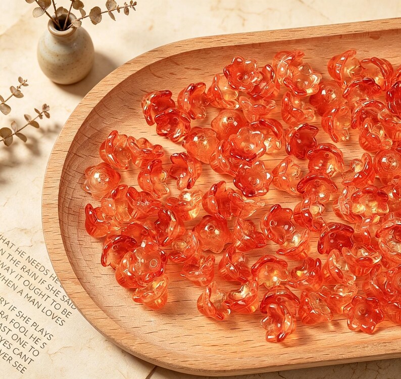 May include: A collection of small, translucent orange flower-shaped beads. Each bead features a five-petal design with a central hole for stringing. The beads are scattered across a smooth, light-colored wooden tray with a curved edge.