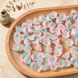 May include: A collection of small, translucent flower-shaped beads in soft shades of pastel pink and mint green. Each bead features a central hole and a five-petaled design, scattered across a smooth, light-colored wooden tray surface.