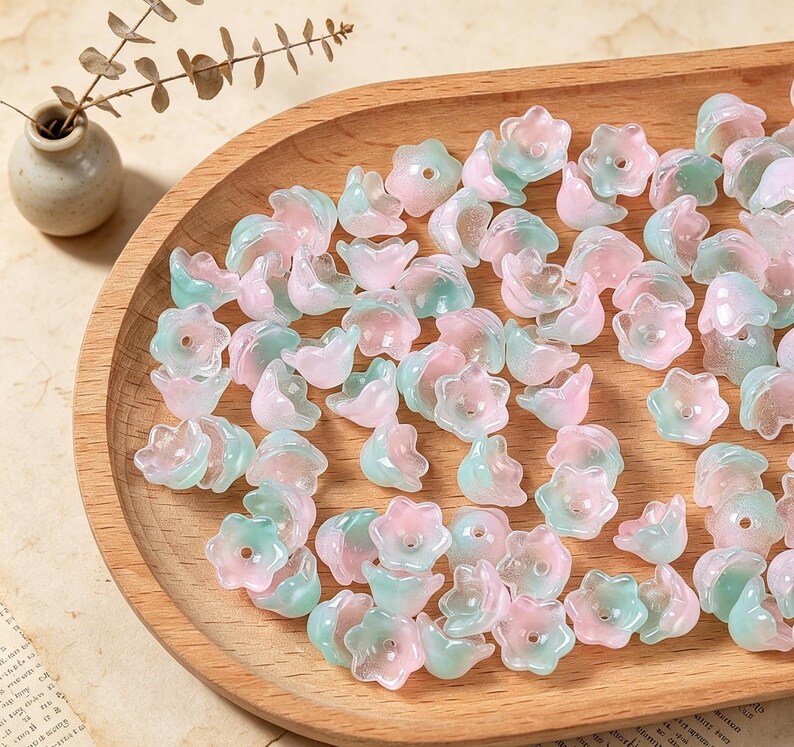 May include: A collection of small, translucent flower-shaped beads in soft shades of pastel pink and mint green. Each bead features a central hole and a five-petaled design, scattered across a smooth, light-colored wooden tray surface.