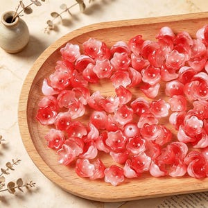May include: A collection of small, translucent flower-shaped beads in varying shades of red, pink, and white. Each bead features a five-petaled design with a central hole, creating a delicate, layered appearance across the wooden surface.