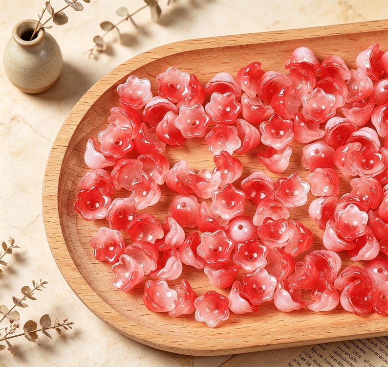 May include: A collection of small, translucent flower-shaped beads in varying shades of red, pink, and white. Each bead features a five-petaled design with a central hole, creating a delicate, layered appearance across the wooden surface.