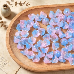 May include: A collection of small, translucent flower-shaped beads in a gradient of soft pink and light blue. Each bead features a central hole and a five-petaled design, scattered across a smooth, light-toned wooden tray with a curved edge.