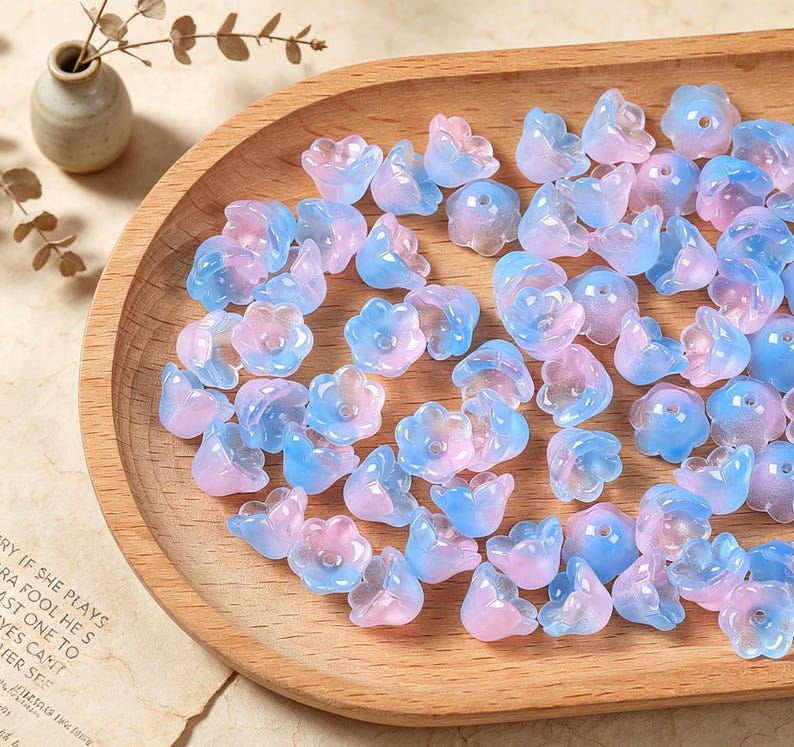 May include: A collection of small, translucent flower-shaped beads in a gradient of soft pink and light blue. Each bead features a central hole and a five-petaled design, scattered across a smooth, light-toned wooden tray with a curved edge.