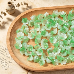 May include: A collection of small, translucent, bell-shaped flower beads in varying shades of light and dark green. Each bead features a central hole for threading, with a smooth, glossy finish that mimics the appearance of delicate lily of the valley.