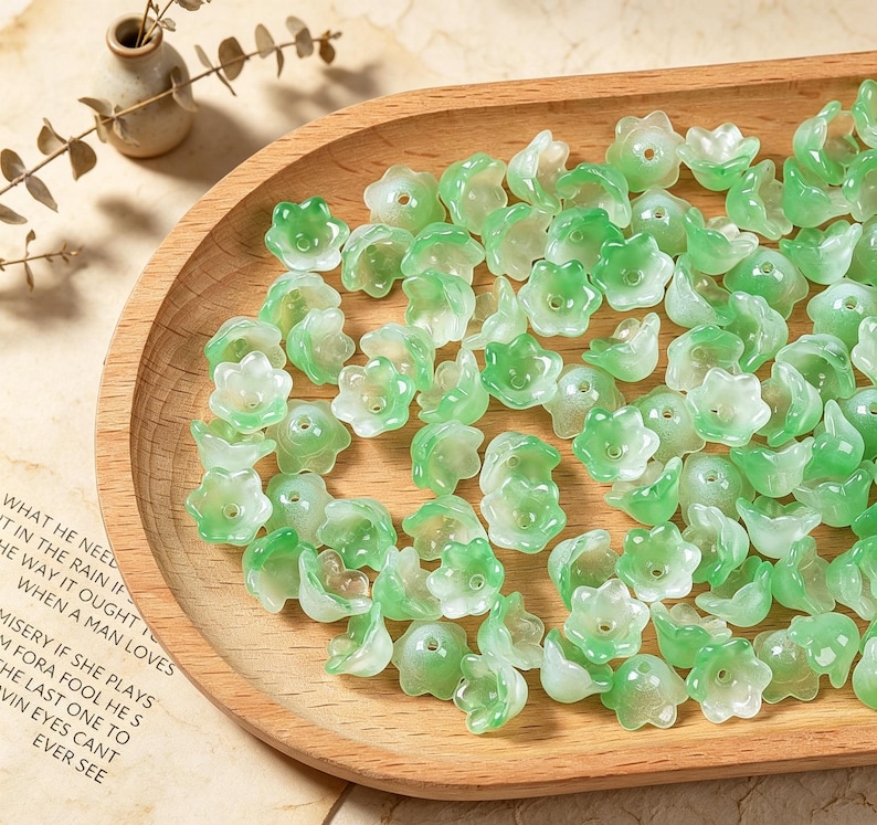 May include: A collection of small, translucent, bell-shaped flower beads in varying shades of light and dark green. Each bead features a central hole for threading, with a smooth, glossy finish that mimics the appearance of delicate lily of the valley.