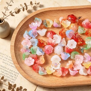 May include: A collection of small, translucent, flower-shaped beads in various colors including pink, blue, red, orange, green, and white. Each bead features a central hole and a five-petaled design, scattered across a smooth, light-toned wooden tray.