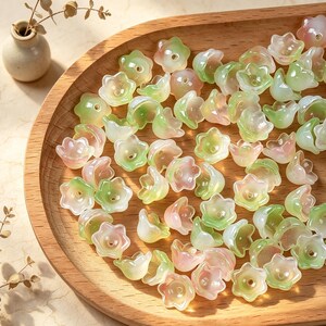 May include: A collection of small, translucent flower-shaped beads in soft shades of pale green and light pink. Each bead features a five-petaled design with a central hole, scattered across a smooth, light-colored wooden tray surface.