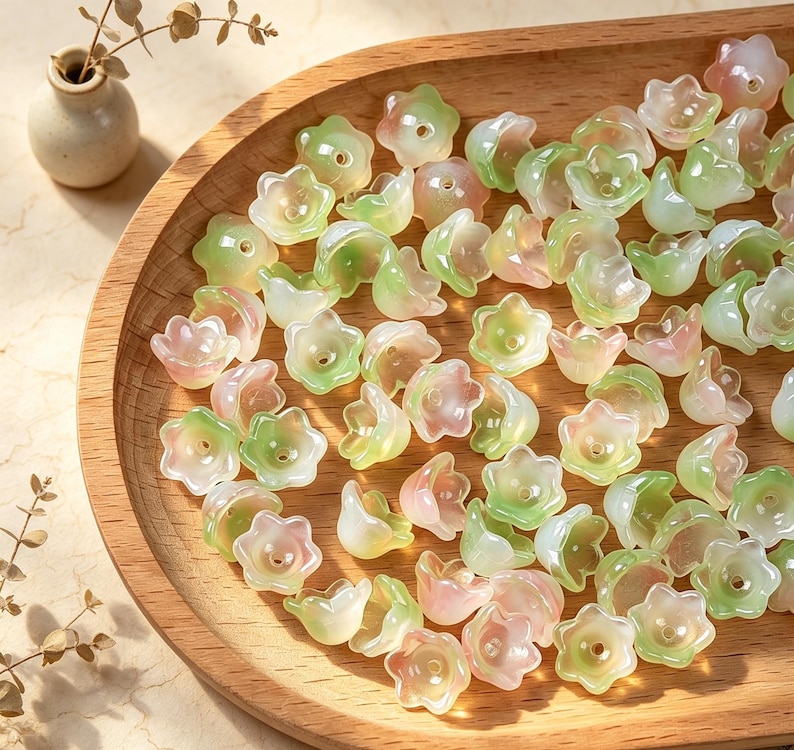 May include: A collection of small, translucent flower-shaped beads in soft shades of pale green and light pink. Each bead features a five-petaled design with a central hole, scattered across a smooth, light-colored wooden tray surface.