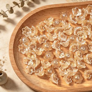 May include: A collection of small, translucent, light amber-colored flower-shaped beads. Each bead features a five-petaled design with a central hole for threading, crafted from a clear material that catches the light to create a subtle glow.