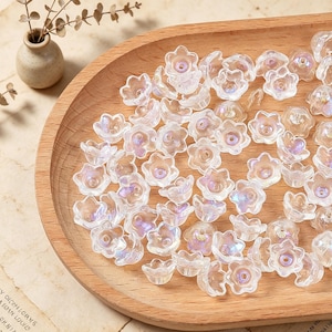 May include: A collection of small, clear, flower-shaped beads with an iridescent finish. Each bead features five petals and a central hole for threading. The beads are scattered across a smooth, light-colored wooden tray with a curved edge.