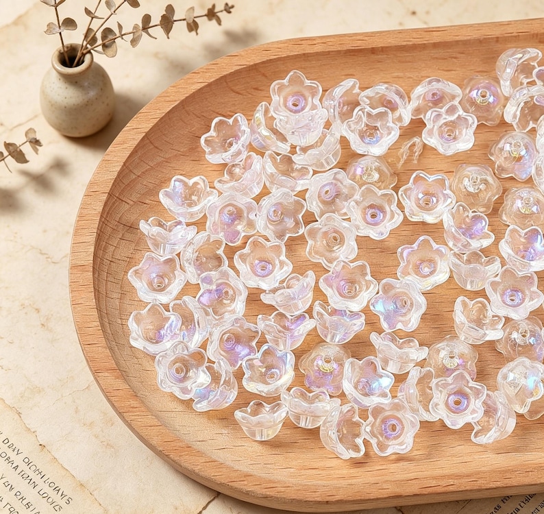 May include: A collection of small, clear, flower-shaped beads with an iridescent finish. Each bead features five petals and a central hole for threading. The beads are scattered across a smooth, light-colored wooden tray with a curved edge.