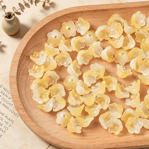 May include: A collection of small, translucent, pale yellow flower-shaped beads with a central hole. Each bead features five rounded petals and a smooth, glossy finish, arranged in a scattered pile on a curved wooden surface.