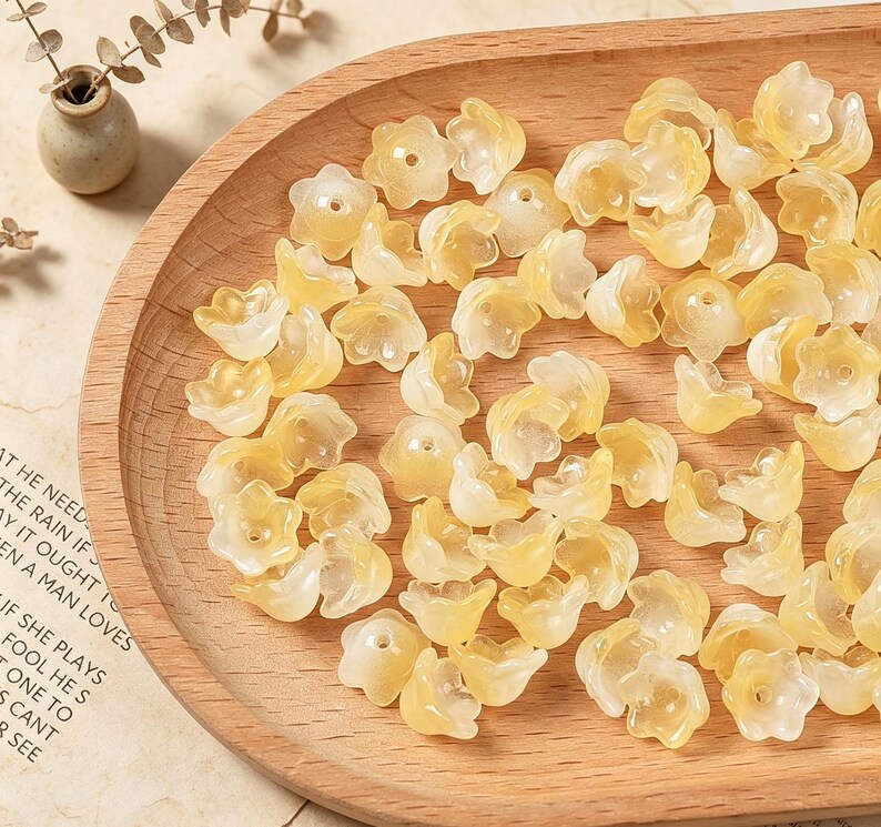 May include: A collection of small, translucent, pale yellow flower-shaped beads with a central hole. Each bead features five rounded petals and a smooth, glossy finish, arranged in a scattered pile on a curved wooden surface.