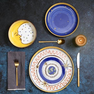 May include: A table setting with celestial designs. It includes plates with moon and star patterns, a gold spoon, fork, and knife, a candle, and a grey napkin. The plates are blue, yellow, and white with gold rims.