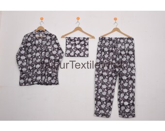 May include: A three-piece pajama set featuring a button-down shirt, a matching scarf, and pants. The set is adorned with a black and white floral pattern. The garments are displayed on wooden hangers against a white background.