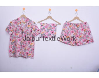 May include: A pink floral pajama set, including a button-down shirt, shorts, and matching shorts, hangs on wooden hangers. The set features a white background with pink and orange flower patterns. The text "Jaipur Textile Work" is visible.