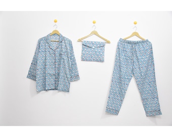 May include: A light blue pajama set with a repeating pattern of white and pink shapes. The set includes a button-down shirt, drawstring pants, and a matching pouch, all hanging on wooden hangers against a white wall.