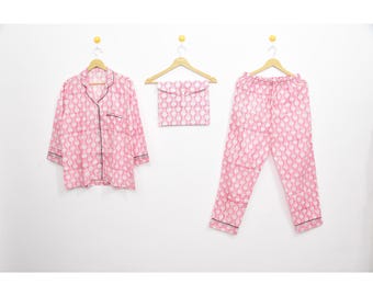 May include: Pink and white patterned pajama set, including a button-down shirt, pants, and a matching pouch. The pajamas have black trim and are hanging on wooden hangers against a white background. The pattern is a repeating design.