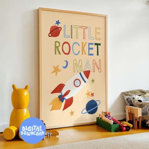 Peut inclure: Une affiche encadrée sur fond beige clair représente une fusée, des planètes et des étoiles. Les mots "LITTLE ROCKET MAN" sont écrits en lettres colorées. Un jouet en forme d'ours jaune et d'autres jouets sont au premier plan.