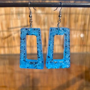 May include: A pair of blue resin earrings with a rectangular shape and a hollow center. The earrings are filled with silver glitter and have silver-colored ear wires. The earrings are hanging from a dark metal bar.
