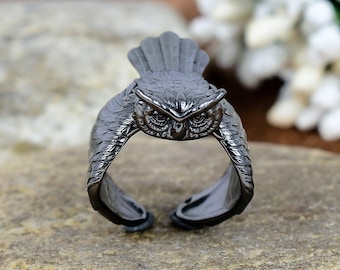 Könnte beinhalten: Ein dunkelgrauer Eulenring. Der Ring hat ein detailliertes Eulen-Design mit strukturierten Federn und großen Augen. Der Ring ist unten offen, was eine verstellbare Passform ermöglicht. Der Eulen-Kopf befindet sich oben, wobei sich die Flügel zu den Seiten erstrecken.