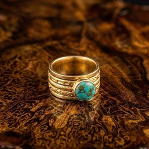 Peut inclure: Une bague en or ornée d'une pierre turquoise. La bague présente un motif gravé sur le pourtour. La pierre turquoise est ronde et sertie dans une lunette en or. La bague est présentée sur une surface marron à motifs.