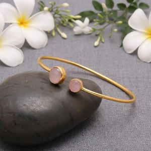 May include: A gold-colored cuff bracelet with two round, light pink gemstones. The bracelet is resting on a dark gray stone, with white flowers and green leaves in the background. The gemstones are set in gold bezels.