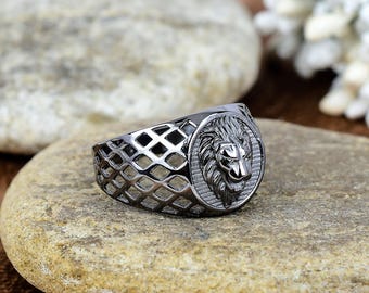 Könnte beinhalten: Ein silberfarbener Ring mit einem Löwenkopf-Design. Der Ring hat einen detaillierten Löwenkopf auf einem kreisförmigen Emblem und ein Gittermuster auf dem Band. Der Ring wird auf einem hellbraunen Stein präsentiert.