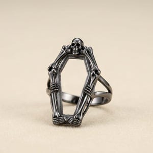 May include: A black ring featuring a coffin-shaped design with a skeleton holding the coffin. The skeleton's skull is at the top of the coffin. The ring is made of a dark metal and is set against a neutral background.