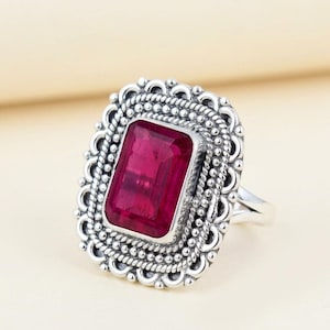 May include: A silver ring with a large, rectangular, deep pink gemstone. The gemstone is set in an ornate frame with a beaded border. The ring's band is a simple, polished silver.