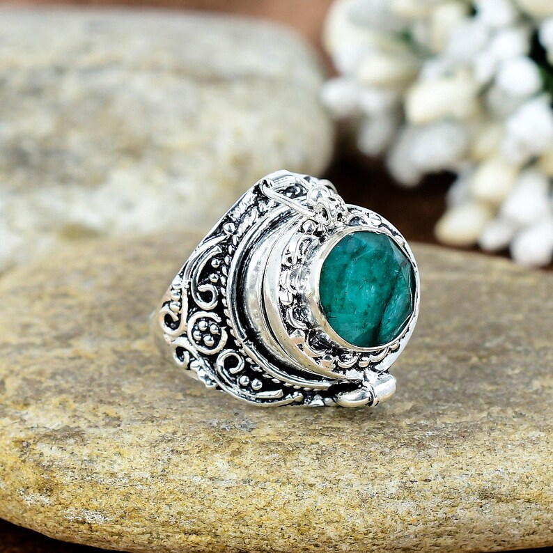 May include: A silver ring with an oval, emerald-green gemstone. The ring has an ornate, detailed design with scrollwork and a hinged compartment. The ring is set on a light brown stone surface.