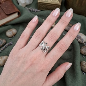 May include: A silver dragon ring with detailed wings and a small gemstone. The ring is worn on a finger, showcasing the intricate design and craftsmanship. The dragon is facing to the right.