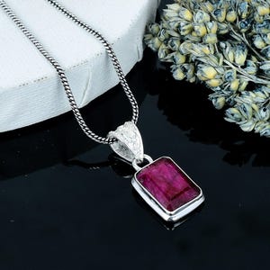 Peut inclure: Un collier pendentif en argent avec une pierre précieuse rectangulaire rouge foncé. La pierre est sertie dans un cadre en argent et suspendue à une fine chaîne en argent. La bélière du pendentif a un design orné.