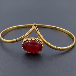 Op de afbeelding: Een goudkleurige metalen armband met een V-vormig middenstuk. Het middelpunt is een ovale, dieprode cabochonsteen die is gezet in een decoratieve, geschulpte metalen rand die de twee zijden van de V-vormige structuur met elkaar verbindt.