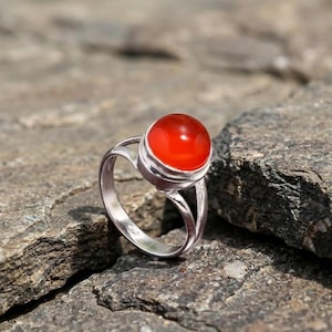 Op de afbeelding: Een zilveren ring met een grote, ronde, helderrode edelsteen. De ring is geplaatst op een getextureerd grijs rots oppervlak. De edelsteen is het middelpunt, vangt het licht en vormt een levendig contrast met de zilveren band.