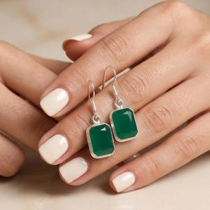 May include: A pair of silver earrings with rectangular, emerald-green gemstones. The earrings are displayed against a neutral background, showcasing their polished metal and vibrant color. The gemstones are faceted, catching the light and adding sparkle.