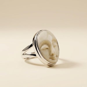 Può includere: Un anello d'argento con un volto scolpito con gli occhi chiusi. Il volto è di colore beige chiaro ed è incastonato in una cornice d'argento. Il gambo dell'anello si divide in due parti su entrambi i lati del volto.
