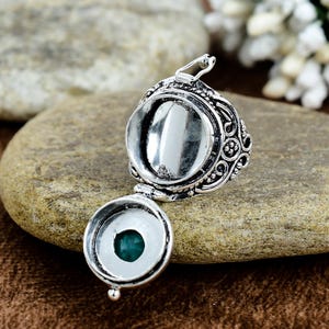 May include: A silver-colored locket with an open compartment and a hinged bottom, revealing a small, round, teal-colored stone. The locket features ornate, swirling patterns and is displayed on a textured, brown stone surface.