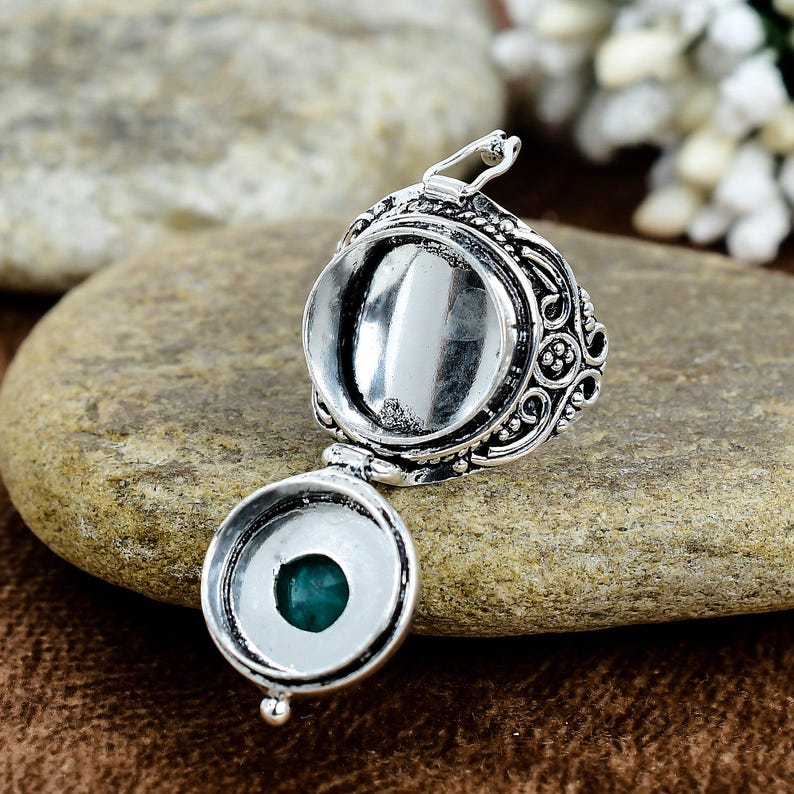 May include: A silver-colored locket with an open compartment and a hinged bottom, revealing a small, round, teal-colored stone. The locket features ornate, swirling patterns and is displayed on a textured, brown stone surface.