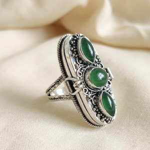 Peut inclure: Bague en argent avec un design ovale allongé, ornée de trois pierres vertes de forme ovale. La bague présente des détails complexes autour des pierres et le long de la bande, avec une bordure texturée en forme de corde.